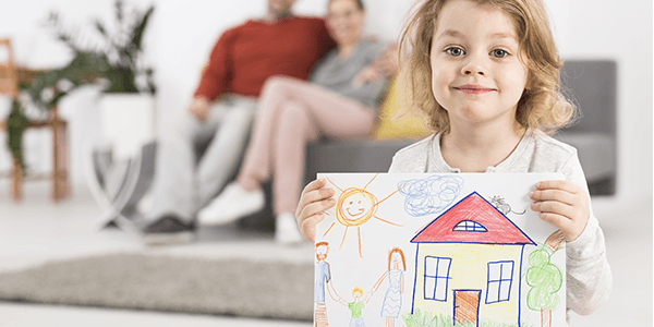 Picture of a little girl holding an drawing of a house with her parents out of focus in the background smiling at her as they sit on a sofa.